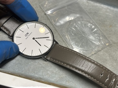 青いゴム手袋をはめた職人の手元。交換が完了した時計の横に、ジップ袋に入った古いガラスの破片がある。