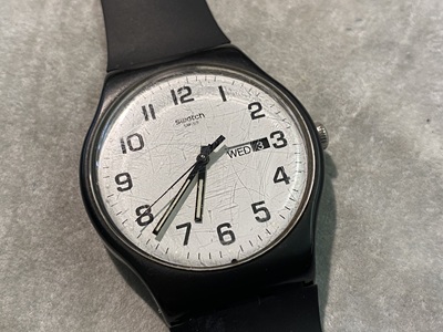 白い文字盤に黒い数字が並ぶ、デイデイト機能付きのスウォッチ腕時計。