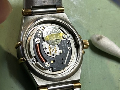 電池が収まっていた金属の接触端子（コンタクトバネ）や、裏蓋の周辺に溜まっていた微細な汚れを、特殊なツールや布できれいに拭き取っている様子。