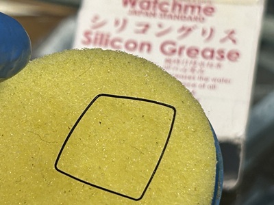 時計メンテナンス用のシリコングリース塗布器の黄色いスポンジの上に、四角いパッキンを載せて均一にグリースを塗布している様子