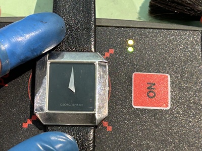 裏蓋を閉じたGEORG JENSEN腕時計をパルスチェッカーの上に載せ、回路から発せられる電子信号の正常点滅ランプを確認している様子