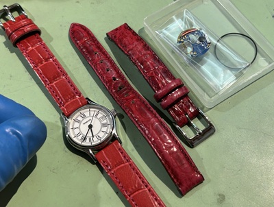 機械が綺麗に生き返ったティファニーの腕時計の横に、これまで使用していた古い赤ベルトと、新しく取り付けるための艶やかで鮮やかな赤色の新品レザーベルトが並べられている様子