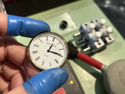 青い指サックをつけた手で、新品ムーブメント交換後に針が正しくセットされたウェッジウッドの時計正面を保持し、動作を確認している職人の手元