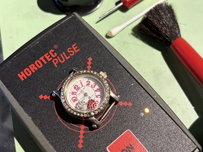 ホロテック（HOROTEC）製のパルスチェッカー機の上に置かれ、正常動作のインジケーターランプが灯る腕時計
