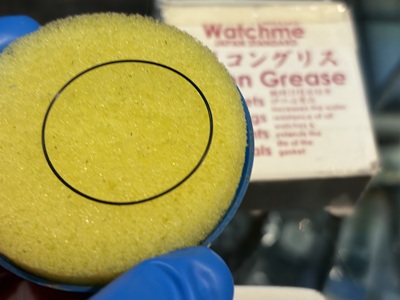 青い指サックをはめた手で、黄色い丸型のシリコングリース塗布器(スポンジ)を持ち、その上に丸い黒い防水パッキンが乗せられているクローズアップ写真。背景には「Watchme シリコングリス Silicon Grease」の文字が見えるパッケージ。