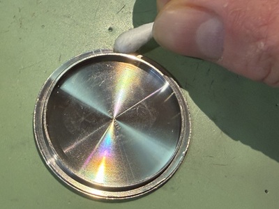 グリーンの作業台の上に置かれた、金属製の丸い裏蓋のフチの隙間や溝を、白い綿棒を使って汚れを優しく拭き取っている手元のクローズアップ写真。