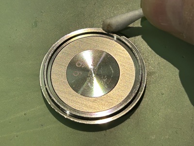外された丸い金属製の裏蓋の内部。外周にある細い防水パッキンが収まる溝のフチを、白い綿棒を使って細やかに拭き掃除し、汗の結晶や汚れを取り除いている手元のクローズアップ写真。