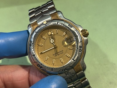 青い指サックをはめた手で持たれた、タグホイヤー(TAG Heuer)プロフェッショナル200のクォーツ式腕時計。ゴールドの文字盤にメルセデス針、3時位置に拡大レンズ付きの日付窓が配置され、ステンレスとゴールドのコンビカラーベゼルが特徴的な時計の画像。