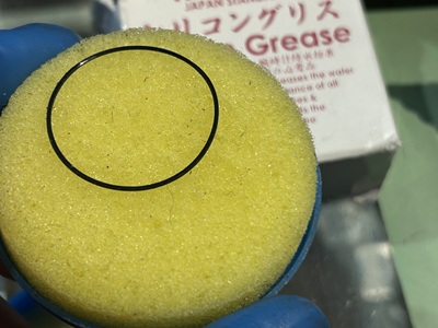 黄色い丸型のパッキン専用グリース塗布器(スポンジ)の上に、黒い防水ゴムパッキンが円形にセットされ、シリコングリースを均一に馴染ませているクローズアップ写真。背景にはグリースのパッケージが見えている。