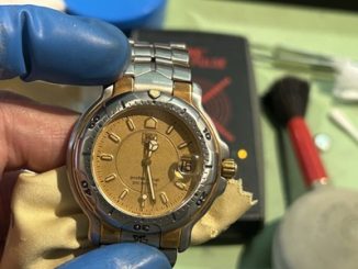 電池交換とすべてのメンテナンスが完了し、正常に動き出したタグホイヤーの腕時計。青い指サックをはめた手で優しく持たれており、背景にはパルスチェッカーや赤い持ち手のブラシが写っている写真。