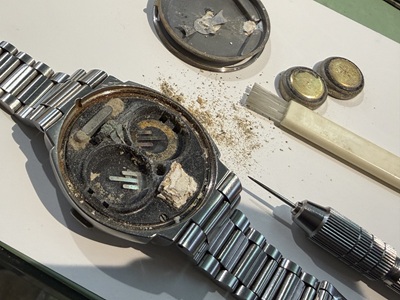 古いボタン電池を取り外し、刷毛や専用工具で内部の粉塵や汚れを掃除している作業風景